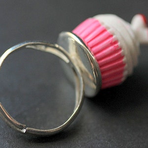 May include: A silver ring with a pink and white cupcake charm. The cupcake has a pink frosting and white sprinkles.