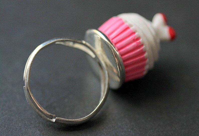 May include: A silver ring with a pink and white cupcake charm. The cupcake has a pink frosting and white sprinkles.