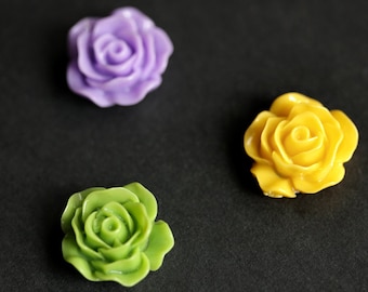 Può includere: Tre abbellimenti a forma di rosa in resina, nei toni del viola, giallo e verde. I fiori sono disposti su uno sfondo scuro, mostrando la struttura dettagliata dei petali. Ideali per progetti di artigianato.
