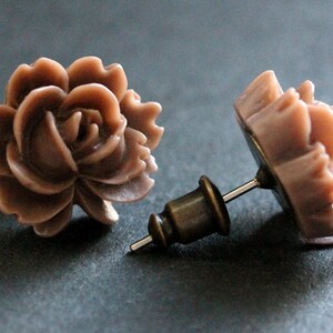 May include: A pair of rose-shaped stud earrings with a brown, almost mauve color. The earrings are made of a smooth, slightly textured material and have a simple post design.
