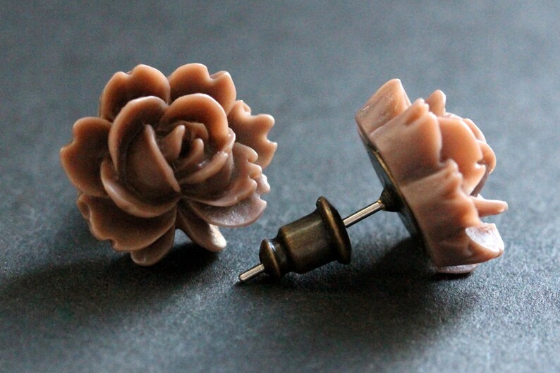 May include: A pair of rose-shaped stud earrings with a brown, almost mauve color. The earrings are made of a smooth, slightly textured material and have a simple post design.