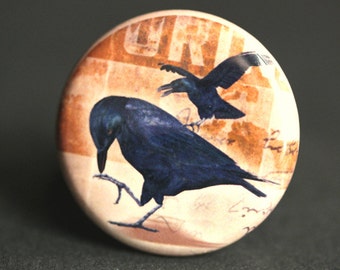 Op de afbeelding: Een ronde knoop met een vintage ontwerp met twee zwarte kraaien op een achtergrond van vervaagd bruin en wit papier met het woord "URI" erop gedrukt.