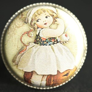 Könnte beinhalten: Eine Brosche im Vintage-Stil mit einem jungen Mädchen in einem weißen Kleid mit Blumenmuster. Das Mädchen hält ein kleines weißes Kaninchen und ist von einem dekorativen Rand umgeben.
