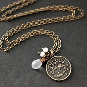 May include: A gold chain necklace with a bronze pendant featuring the astrological symbol for Pisces. The pendant has a white glass bead attached to it.