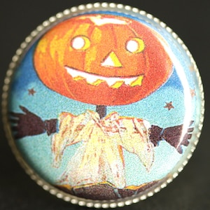 May include: A round, silver-rimmed pin with a vintage Halloween image. A smiling jack-o'-lantern with a white face and black eyes is wearing a white and yellow costume and has black arms outstretched against a blue background with white stars.