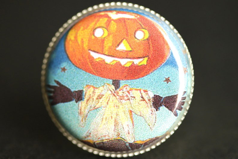 May include: A round, silver-rimmed pin with a vintage Halloween image. A smiling jack-o'-lantern with a white face and black eyes is wearing a white and yellow costume and has black arms outstretched against a blue background with white stars.