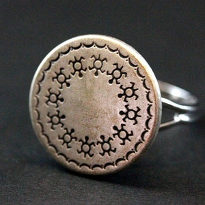 May include: A silver ring with a round, decorative top featuring a repeating pattern of stylized sunbursts. The ring is open and adjustable.