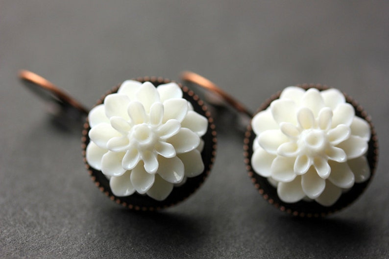 May include: A pair of white flower earrings with copper-toned settings. The flowers are made of white ceramic and have a delicate, intricate design.