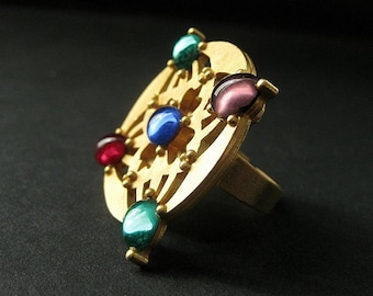 Puede incluir: Un anillo de oro con un diseño decorativo que presenta cinco piedras preciosas de colores. El anillo tiene una banda ancha y una parte superior circular con una piedra preciosa azul central rodeada de una piedra preciosa verde, roja y dos piedras preciosas moradas.