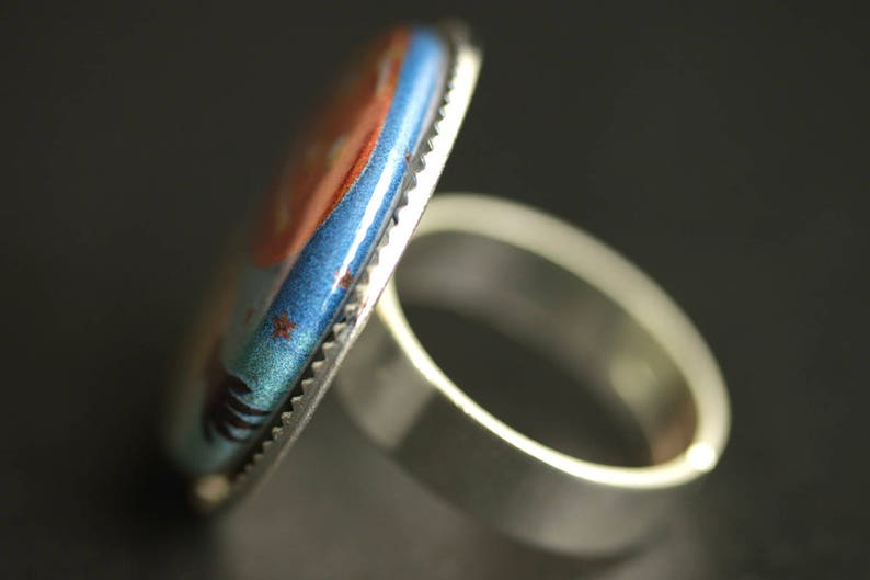 May include: A silver ring with a blue and orange enamel design. The ring has a wide band and a raised design.