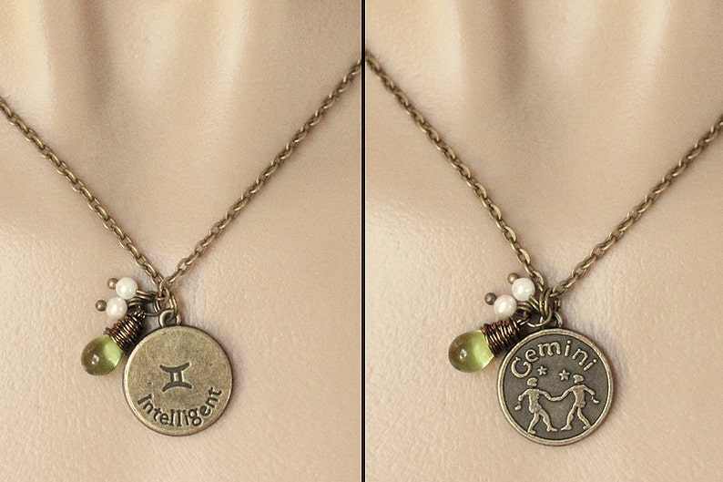 May include: A gold necklace with a round pendant featuring the Gemini zodiac symbol and the word "Intelligent". The pendant is accented with green and white beads.