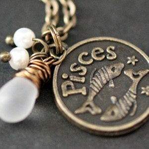 May include: A silver necklace with a charm featuring the zodiac sign Pisces. The charm is a round coin with the word "Pisces" and two fish. The necklace also has a small white bead charm.