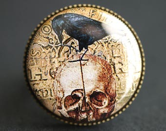 Op de afbeelding: Een ronde, goudkleurige metalen ring met een glazen cabochon met daarop een zwarte raaf die op een schedel met een kruis zit. De schedel is omgeven door ingewikkelde, gedetailleerde patronen en letters.