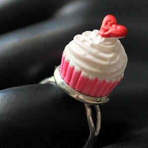 May include: A silver ring with a pink and white cupcake charm. The cupcake has white frosting and a red heart on top.