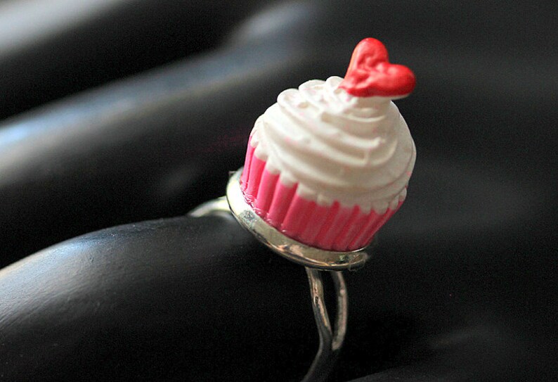 May include: A silver ring with a pink and white cupcake charm. The cupcake has white frosting and a red heart on top.