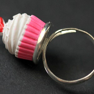 May include: A silver ring with a pink and white cupcake charm. The cupcake has a red heart on top.