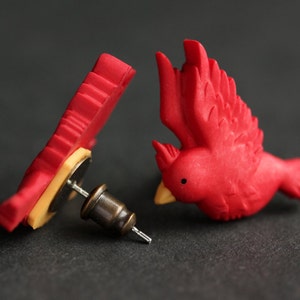 Puede incluir: Un par de pendientes de cardenal rojo. Los pendientes est&aacute;n hechos de arcilla polim&eacute;rica y tienen un dise&ntilde;o realista. Los pendientes est&aacute;n unidos a postes de plata.