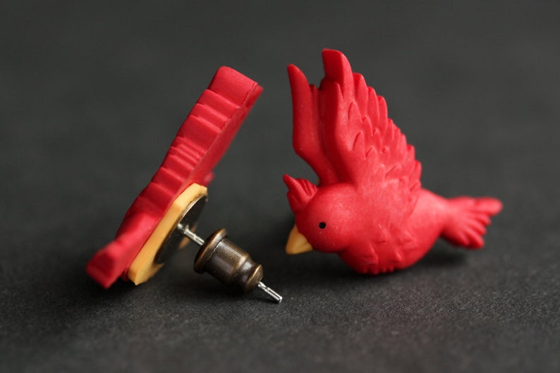 Puede incluir: Un par de pendientes de cardenal rojo. Los pendientes est&aacute;n hechos de arcilla polim&eacute;rica y tienen un dise&ntilde;o realista. Los pendientes est&aacute;n unidos a postes de plata.