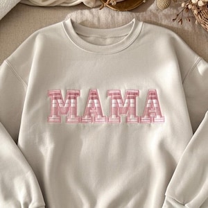 May include: A light beige sweatshirt with the word "MAMA" in pink, block letters across the chest. The letters have a textured, dimensional appearance. The sweatshirt has long sleeves and a crew neck.