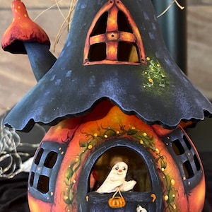 Op de afbeelding: Een decoratief kalebashuisje geschilderd in oranje en zwart met een donkerblauw dak, voorzien van een uitgesneden raam met een geschilderde geest die een pompoenemmer vasthoudt, een kleine witte hond, een schoorsteen met een rode paddenstoel.