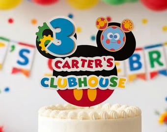 May include: A two-tier white frosted cake decorated with colorful sprinkles and a custom cake topper featuring a black mouse-ear silhouette, the number 3, the text Carter's Clubhouse, and a small blue character face with red and yellow accents.