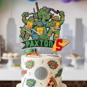 May include: A two-tier white cake decorated with Teenage Mutant Ninja Turtles imagery, including a custom topper with the name Paxton and the number 5. The cake features comic-style sound effects, pizza slices, and brick patterns, surrounded by pizza boxes.