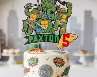 May include: A two-tier white cake decorated with Teenage Mutant Ninja Turtles imagery, including a custom topper with the name Paxton and the number 5. The cake features comic-style sound effects, pizza slices, and brick patterns, surrounded by pizza boxes.