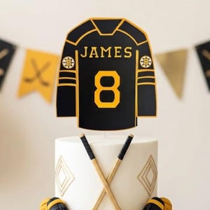 May include: A two-tier white cake topped with a black and gold hockey jersey cutout featuring the name JAMES and number 8. The cake is decorated with crossed hockey sticks, black gloves, and hockey pucks, surrounded by hockey-themed party decor.