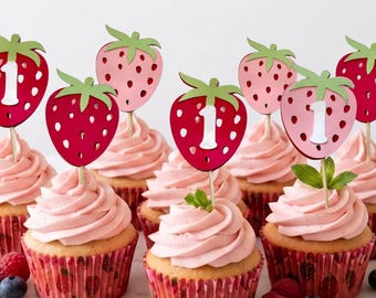 May include: A collection of cupcakes topped with pink frosting and strawberry-shaped cake toppers. Some toppers feature the number one in white. The cupcakes are in red patterned liners and arranged on a white scalloped platter with fresh berries.