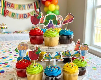 May include: A tiered cupcake stand holds cupcakes topped with colorful frosting and paper toppers featuring illustrations of a green caterpillar, red apple, slice of pie, and butterfly. The toppers are inspired by The Very Hungry Caterpillar.