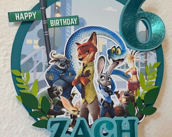 Op de afbeelding: Een Zootopia-thema verjaardagstaarttopper met de personages Nick Wilde en Judy Hopps. De topper bevat de naam "ZACH" en het nummer "6", met de woorden "HAPPY BIRTHDAY" op een straatnaambord.