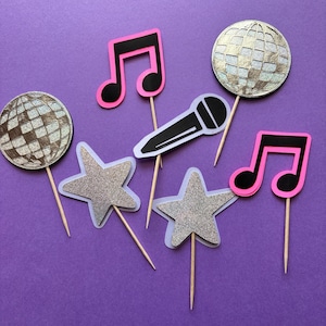 May include: A collection of party decorations on a purple background. The decorations include silver glitter stars, disco ball shapes, black and pink musical notes, and a microphone. Each decoration is attached to a wooden stick.