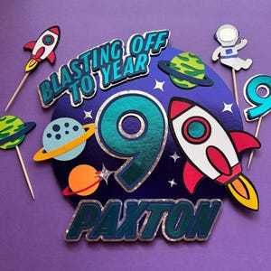 May include: A colorful cake topper with a space theme. It features a large number 9, rockets, planets, and the text "Blasting Off To Year" and "Paxton". The design is primarily blue, red, and green, with silver accents.