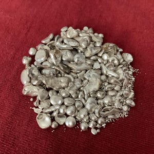 May include: A pile of irregularly shaped, silver-colored metal pieces, possibly ingots or scrap, rests on a deep red fabric. The pieces vary in size and shape, with some appearing smooth and rounded, while others are more jagged.