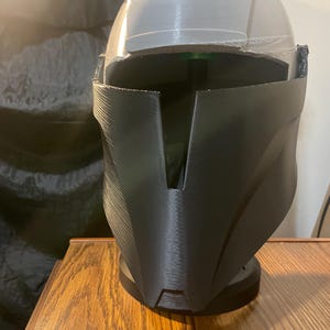 May include: A black, futuristic-style helmet with a visor and a stand. The helmet has a smooth, matte finish and a distinctive angular design. The visor is a light gray color. The helmet is sitting on a wooden surface.