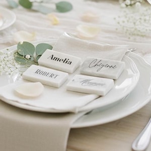 May include: A beautifully set table with white plates, napkins, and glassware. Each place setting includes a name card with a guest's name. A small roll sits on a side plate. The table is decorated with greenery and flower petals.