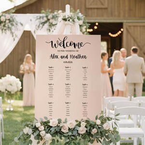 May include: A wedding seating chart on a white easel, with the names "Alex and Heather" and a list of names. The sign is decorated with flowers and draped fabric. The background features a barn and wedding guests.