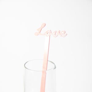 May include: A pink drink stirrer with the word "Love" in cursive script. The stirrer is placed inside a clear champagne flute, set against a white background. The overall theme is romance and celebration.