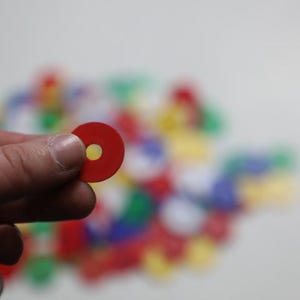 K&ouml;nnte beinhalten: Eine Hand h&auml;lt ein rotes, kreisf&ouml;rmiges Spielteil mit einem gelben Loch in der Mitte. Ein verschwommener Hintergrund zeigt eine Sammlung &auml;hnlicher Spielteile in verschiedenen Farben, darunter Rot, Gr&uuml;n, Blau und Gelb. Die Spielteile scheinen aus Kunststoff zu sein.