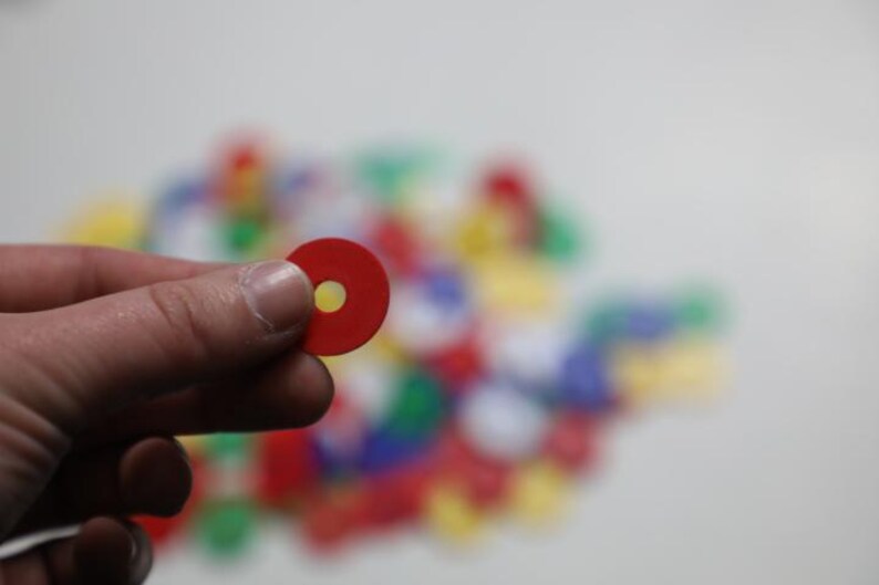 K&ouml;nnte beinhalten: Eine Hand h&auml;lt ein rotes, kreisf&ouml;rmiges Spielteil mit einem gelben Loch in der Mitte. Ein verschwommener Hintergrund zeigt eine Sammlung &auml;hnlicher Spielteile in verschiedenen Farben, darunter Rot, Gr&uuml;n, Blau und Gelb. Die Spielteile scheinen aus Kunststoff zu sein.