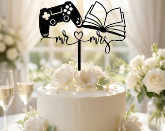 Op de afbeelding: Een zwarte taarttopper met een videogamecontroller aan de linkerkant en een open boek aan de rechterkant, verbonden door de tekst "mr & mrs" met een hartsymbool in het midden. De topper is bevestigd op een enkele verticale prikker.