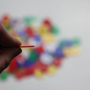 K&ouml;nnte beinhalten: Eine Hand h&auml;lt ein d&uuml;nnes, rotes Spielteil aus Kunststoff. Das Teil wird zwischen Daumen und Zeigefinger gehalten. Ein verschwommener Hintergrund zeigt eine Sammlung bunter Spielteile in Rot, Gr&uuml;n, Blau und Gelb.