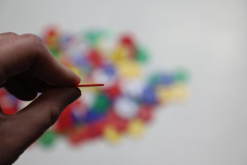 K&ouml;nnte beinhalten: Eine Hand h&auml;lt ein d&uuml;nnes, rotes Spielteil aus Kunststoff. Das Teil wird zwischen Daumen und Zeigefinger gehalten. Ein verschwommener Hintergrund zeigt eine Sammlung bunter Spielteile in Rot, Gr&uuml;n, Blau und Gelb.