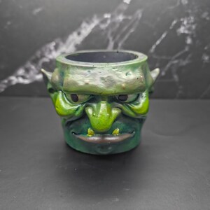 May include: A green goblin-faced planter with a wide open mouth and pointed ears. The planter is painted in shades of green and brown, with a dark interior. The planter is sitting on a dark surface.