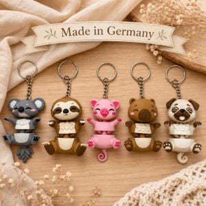 Könnte beinhalten: Fünf tierförmige Schlüsselanhänger in Form eines Koalas, Faultiers, Schweins, Bären und Mopses. Jede Figur ist in zwei Abschnitte unterteilt, die durch eine Metallkette und einen Ring verbunden sind. Der Koala ist grau, das Faultier braun, das Schwein rosa, der Bär braun und der Mops beige.