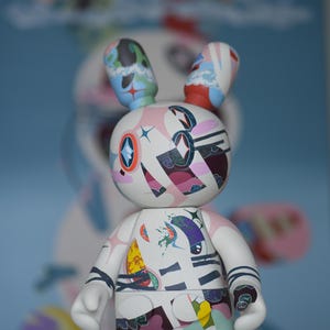 Peut inclure: Figurine de lapin blanc et abstrait avec des motifs et des formes colorées. Le lapin a des accents bleus, verts et rouges sur ses oreilles et son corps. La figurine est posée sur une surface blanche avec un fond bleu flou.