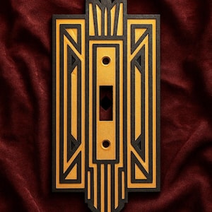 May include: Art Deco style light switch cover in black and gold. The cover features a geometric design with a rectangular shape and a central opening for the switch. The color scheme is a combination of black and gold.