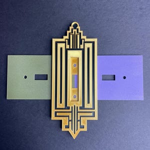 May include: A gold-colored, Art Deco-style light switch cover with a rectangular center and intricate geometric designs. Two rectangular plates, one olive green and one purple, are positioned on either side.