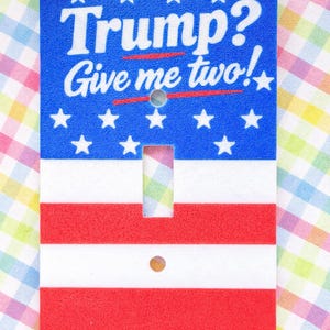 May include: A decorative light switch cover with a patriotic design. The top section is blue with white stars and the text "Trump? Give me two!" The bottom section has red and white stripes.