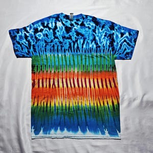 Può includere: Una maglietta tie-dye con una vivace combinazione di colori. La parte superiore presenta un motivo blu e nero, che si trasforma in strisce verticali verdi, arancioni, gialle e blu. La maglietta è realizzata in un materiale morbido.